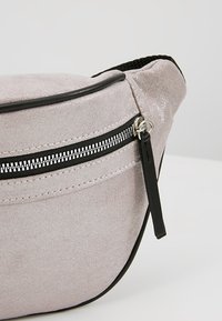 Sac banane en daim gris clair avec fermeture éclair noire, languette noire et bordure noire sur fond blanc uni.