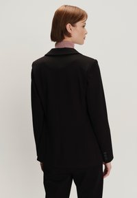 Zwarte op maat gemaakte blazer met een gestructureerd ontwerp, met notch-revers en vier knopen op de manchetten. Zachte stof, minimale details en een getailleerde pasvorm.
