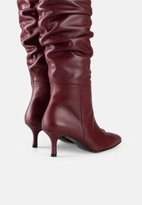 Botine de piele burgundy, cu înălțime până la genunchi, cu vârfuri ascuțite și tocuri subțiri, pe un fundal alb.