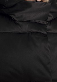 Person som bär en svart vadderad vinterjacka med hög krage som täcker nacken och en del av hakan.