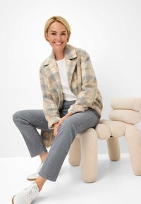Manteau en laine à carreaux beige et bleu avec un col cranté, porté sur un haut blanc, associé à un jean gris et des baskets blanches, assis sur une chaise de couleur claire.