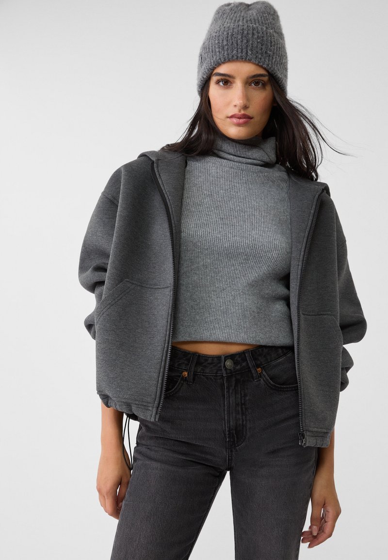 Donna che indossa un berretto grigio a maglia, un dolcevita grigio a coste, una felpa con zip scura e jeans neri a vita alta su uno sfondo chiaro e uniforme.
