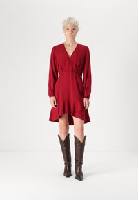 Robe enveloppante rouge avec des manches longues, un ourlet volanté et un décolleté en V profond. Associée à de hautes bottes de cowboy marron avec des coutures décoratives.