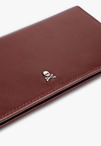 Cartera de cuero color granate con una textura suave, que presenta un emblema plateado de calavera y huesos cruzados. Diseño sencillo con detalles de costura prolija.