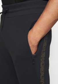 Black sweatpants with a ribbed waistband. Features a zippered pocket and a logo detail in yellow on the side. Smooth fabric texture.