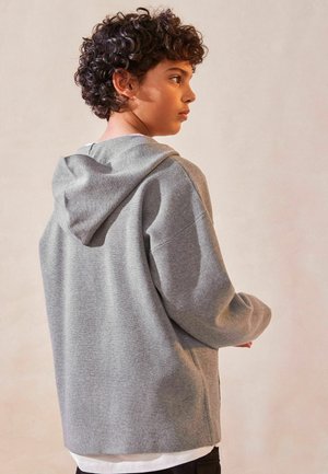 Szary sweatshirt z miękką teksturą, z kapturem, luźnym krojem i zaokrąglonym wykończeniem. Stylizowany na białej koszulce, widok od tyłu.