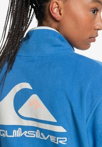 Blue fleece jacket with a high collar, featuring a white logo design with an orange accent. Soft texture, casual style, and relaxed fit.