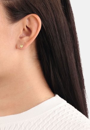 Gold heart-shaped stud earring displayed on an ear. Smooth surface, small size, minimalist design against straight, dark hair and a white top.