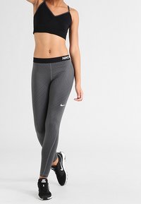 Czarny stanik sportowy i szare legginsy w wzory, oba o dopasowanym kroju. Legginsy mają logo Nike i wysoki pas. Czarne buty sportowe.