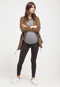 Conjunto de maternidade com uma jaqueta leve castanha, camisa listrada e leggings pretas. O modelo usa tênis brancos.