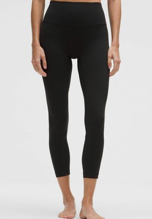 Frau trägt hoch taillierte schwarze, gekürzte Leggings, steht barfuß vor einem schlichten hellgrauen Hintergrund.