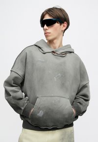 Γκρι υπερμεγέθη φούτερ με ξεθωριασμένο σχέδιο, μπροστινή τσέπη και ακατέργαστες λεπτομέρειες στο στρίφωμα. Συνδυασμένο με μαύρα γυαλιά ηλίου τύπου βισέρ για μια σύγχρονη εμφάνιση.