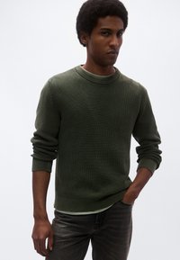 Zeleni pulover z rebrasto teksturo, z okroglim izrezom, teksturirano tkanino in zvitimi manšetami. Nošen preko svetlo zelene srajce, v kombinaciji s temnimi kavbojkami.