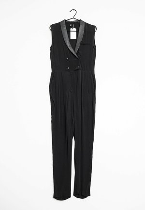 Combinaison sans manches noire avec un col satiné, des boutons double boutonnage, des poches latérales et des jambes fuselées. Accrochée à un crochet noir.