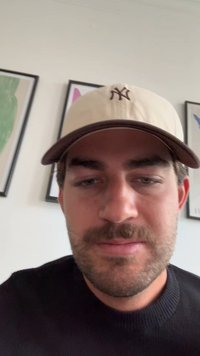 Beige színű baseball sapka sötétbarna karimával, hímzett logóval, amelyet egy fekete pulóveres személy visel, a háttérben világos színű falakkal.