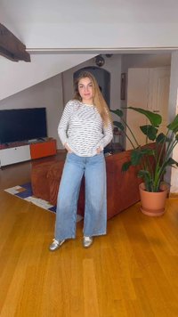 Jonge vrouw staand met handen in de zakken, draagt een gestreept shirt met lange mouwen, wijde jeans, zilveren schoenen, in een woonkamer met planten en tv.