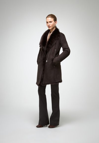 Palton din suede maro cu guler din blană, buzunare laterale cu fermoar și croi drept. Modelul poartă pantaloni gri ajustați și pantofi cu toc.