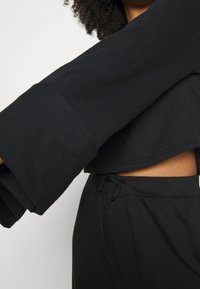 Top corto negro con mangas anchas, hecho de tela suave; combinado con pantalones negros de cintura ajustable con cordón, que muestran un detalle de corte en el abdomen.