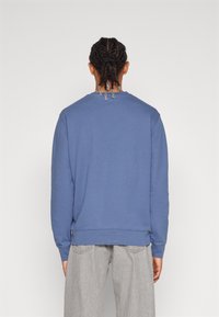 Katoenen sweatshirt in blauw met lange mouwen, ronde hals en geribde manchetten. Heeft een relaxte pasvorm en is gecombineerd met lichtgrijze broeken.