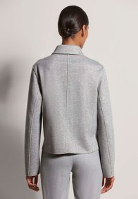 Grijze visgraatjas met een kraag, witte bies langs de naden en lange mouwen, van achteren weergegeven. Vlotte textuur.