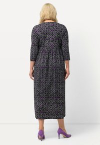 Maxikleid aus schwarzem Stoff mit einem floralen Muster in Lila und Weiß, mit drei Viertelärmeln und einem raffung an der Taille. Kombiniert mit lila High Heels.