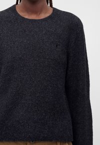 Personne portant un pull à manches longues sombre et duveteux ainsi qu’un pantalon marron, montrée des épaules à la taille sur un fond uni.
