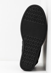 Semelle de chaussure noire avec motifs losange et bandes, taille indiquée 37, positionnée verticalement contre des murs d'angle blancs.