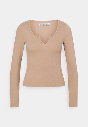 Haut à manches longues en tissu côtelé beige clair, avec un décolleté en V festonné et une silhouette ajustée, agrémenté de détails de coutures minimaux.