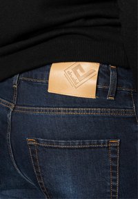 Vaqueros de denim con un lavado azul oscuro, que presentan costuras naranjas y un parche rectangular de cuero marrón claro con un logo en relieve en la cinturilla trasera.