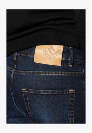 Denim jeans with a dark blue wash, featuring orange stitching and a rectangular, tan leather patch with embossed logo on the back waistband.