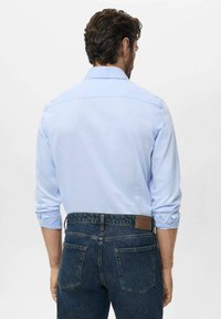 Man gezien van achteren, gekleed in een lichtblauw overhemd met lange mouwen en opgerolde manchetten, en donkerblauwe denimjeans, tegen een effen achtergrond.