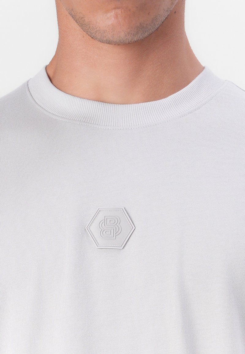 T-shirt grigio chiaro a maniche corte con scollo rotondo. Presenta un logo esagonale testurizzato in un colore grigio chiaro abbinato sul petto.