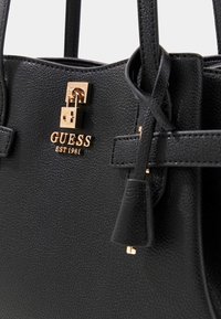 Czarna torebka o teksturowanej powierzchni z złotymi elementami, logo "GUESS" oraz strukturalnym kształtem, wyposażona w dwie rączki i odpinany pasek.