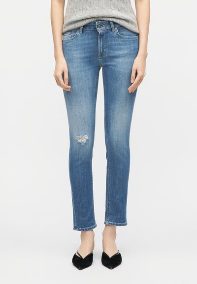 Γυναίκα που φοράει μπλε στενά παντελόνια μέσης μεσαίου ύψους με μικρή σκισμένη λεπτομέρεια στον αριστερό μηρό και μαύρα slingback παπούτσια με μυτερή μύτη.