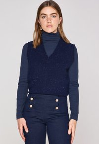 Navyblauem, gestricktem ärmellosem Pullover mit V-Ausschnitt, strukturierter Stoff mit mehrfarbigen Flecken. Kombiniert mit hochgeschnittenen navyblauen Jeans mit goldenen Knöpfen.