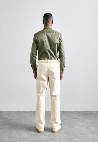 Cămașă verde măsliniu cu mânecă lungă, croi ajustat, asortată cu pantaloni drepți de culoare crem și încălțăminte deschisă, cu talpă texturată.