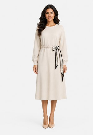 Kvinde iført en beige midi-kjole med lange ærmer, matchende bælte og dekorativ sløjfe, kombineret med beige spidse hæle.