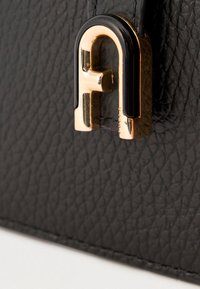 Mustast tekstureeritud nahast kott, millel on silmatorkav kuldse ja musta logoga aksent. Sile viimistlus nähtava õmbluste detailiga.