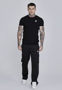 Fekete pamut póló kerek nyakkal és kis logóval a mellkasán, fekete cargo nadrággal párosítva, ami oldalzsebekkel és felhajtott szárakkal rendelkezik.