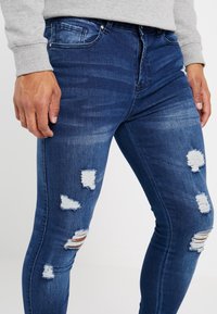 Jeans de denim azul ajustados con áreas desgastadas y bordes deshilachados, cinco bolsillos y un cierre clásico de cremallera y botón.