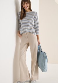 Femme souriante, portant un haut à manches longues rayé, un pantalon large beige, des baskets argentées, et tenant un grand sac fourre-tout bleu clair.