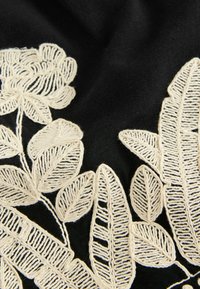 Schwarzer Stoff mit aufwendiger weißer Stickerei von floralen und Blattmustern, die detaillierte Nähte und unterschiedliche Texturen zeigt.