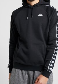 Sudadera negra con capucha, mangas raglán y bolsillo frontal. Presenta un logo blanco en el pecho y franjas laterales blancas con un patrón de logo repetido.