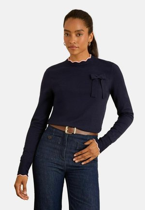 Marineblauer Pullover mit langen Ärmeln, rosa gewelltem Kragen und Bündchen, mit einem kleinen Schleifendetail auf der linken Brust. Getragen mit dunkler Jeans.