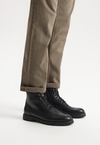 Czarne skórzane botki za kostkę z sznurowanym przodem, gumową podeszwą i zaokrąglonym noskiem, w parze z podwiniętymi khaki spodniami. Gładka tekstura, zorganizowany design.