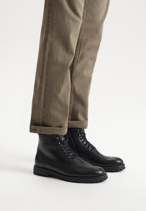 Czarne skórzane botki za kostkę z sznurowanym przodem, gumową podeszwą i zaokrąglonym noskiem, w parze z podwiniętymi khaki spodniami. Gładka tekstura, zorganizowany design.