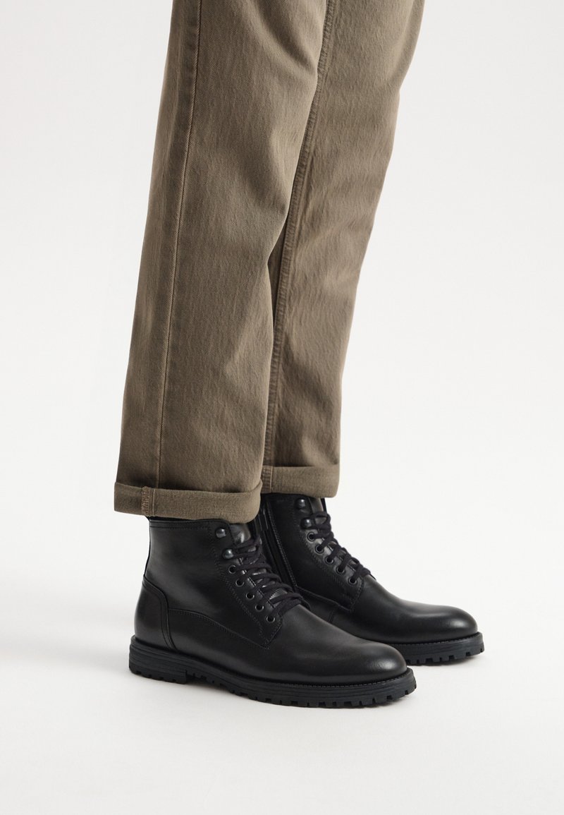 Czarne skórzane botki za kostkę z sznurowanym przodem, gumową podeszwą i zaokrąglonym noskiem, w parze z podwiniętymi khaki spodniami. Gładka tekstura, zorganizowany design.