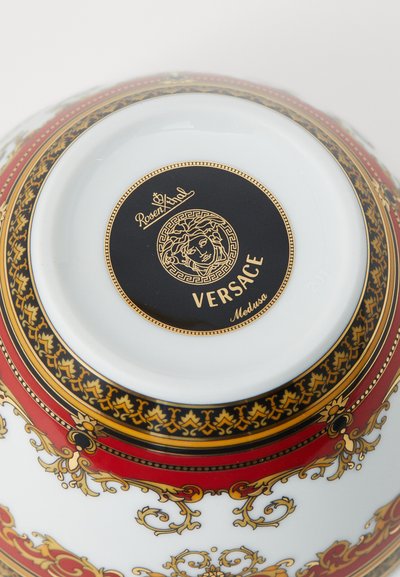 Στρογγυλό πορσελάνινο μπολ με λευκή βάση, περίτεχνα κόκκινα και χρυσά σχέδια και μαύρο λογότυπο Versace στο κάτω μέρος.