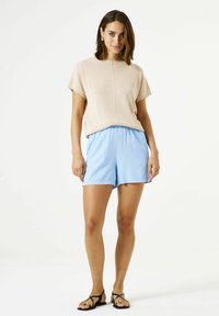 Beige katoenen top met korte mouwen en een ronde hals, gecombineerd met lichtblauwe shorts met elastische taille. Het model draagt zwarte sandalen.