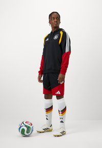 Hoodie atlético preto e vermelho com detalhes em amarelo, combinado com calções pretos com detalhes em vermelho. Meias brancas com as cores da bandeira da Alemanha e chuteiras.